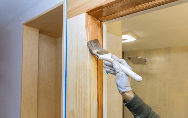 Hands applying varnish on a wooden casement window frame with a brush.