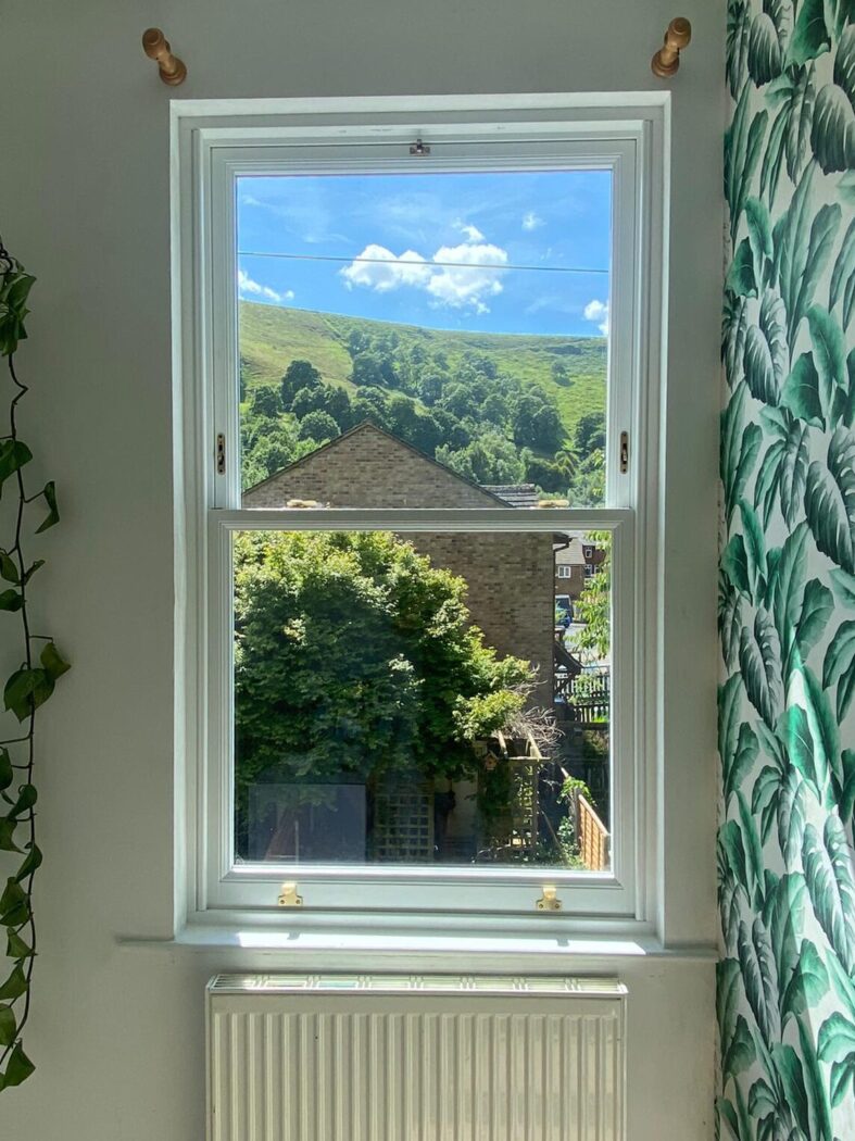 Traditional weights and pulleys sliding sash window in a bedroom with green leaf print wall paper, hanging ivy and a view out over Hebden Bridge on a sunny day