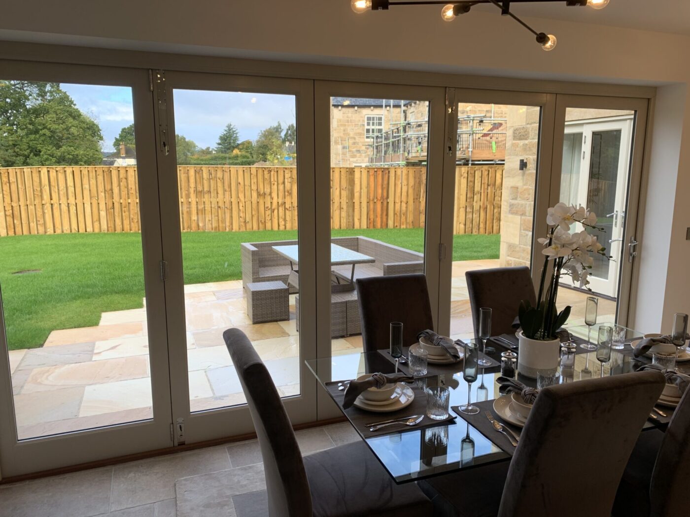 Dining room with a glass table with set places and orchids in the middle. The dining room has a wooden bi fold door which looks out over a patio with garden furniture.