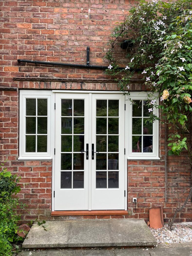 Patio of a red brick house with magloia french doors with georgian bars and black iron handles. The patio has an overhanging tree with lilac flowers.