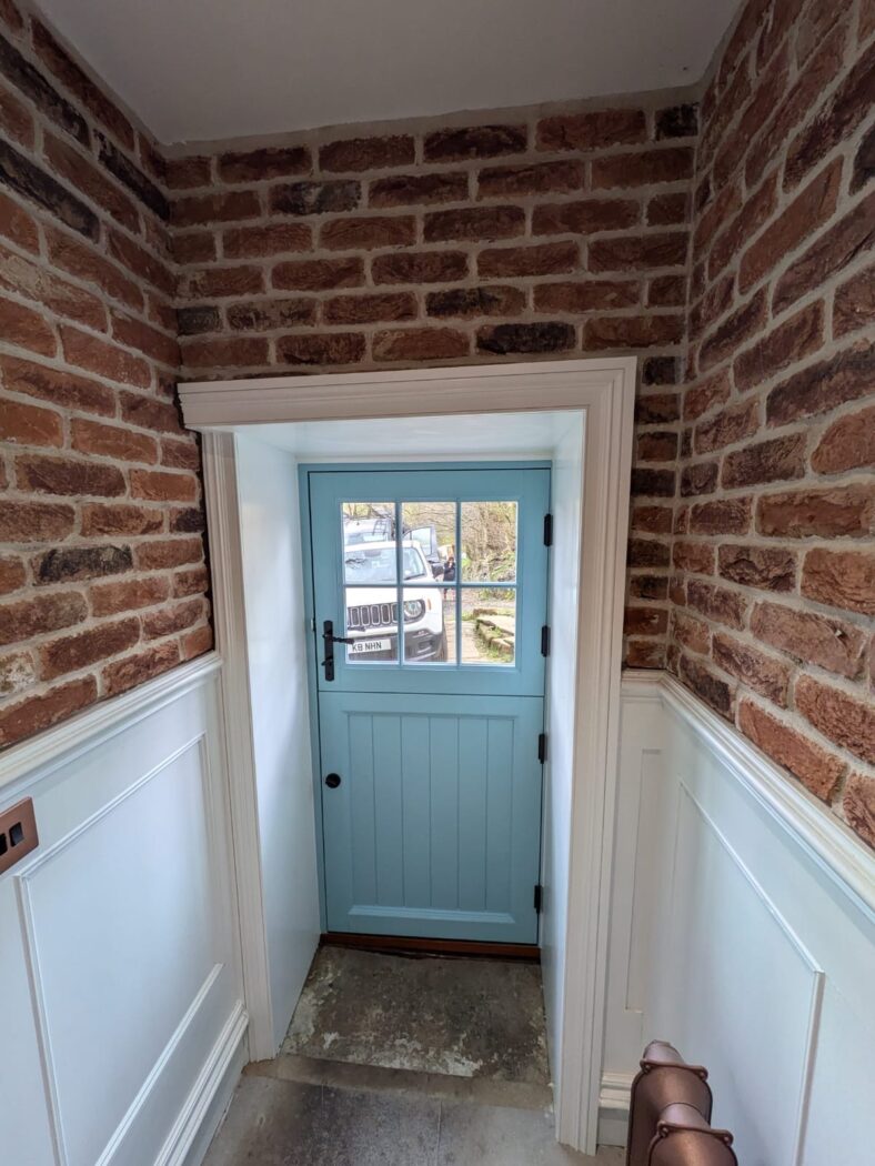 Exposed brick hallway with a new light blue wooden stable door with black iron handle