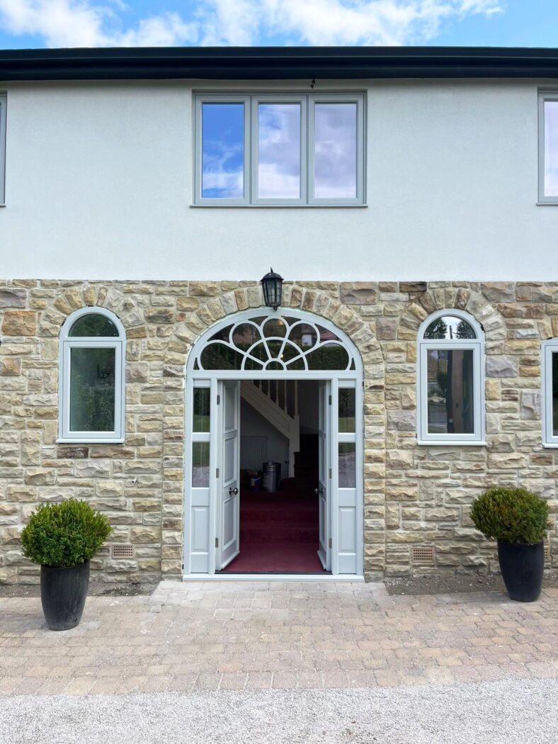 Large house with white wash and stone walls with a set of large double doors opening inwards. Either side of the door is a set of potted plants.