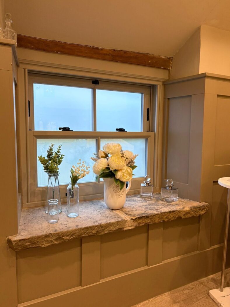 A softly lit bathroom colour drenched in Farrow & Ball Light Grey. A traditional sash window sits above a stone window sill with a jug of large white flowers.